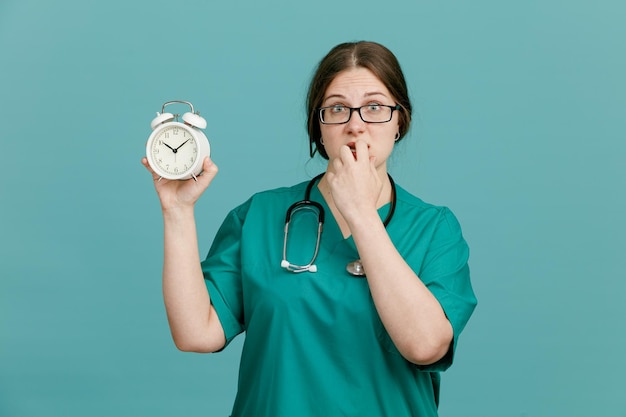 Enfermeira jovem em uniforme médico com estetoscópio no pescoço mostrando despertador olhando para câmera estressado e nervoso roer unhas em pé sobre fundo azul