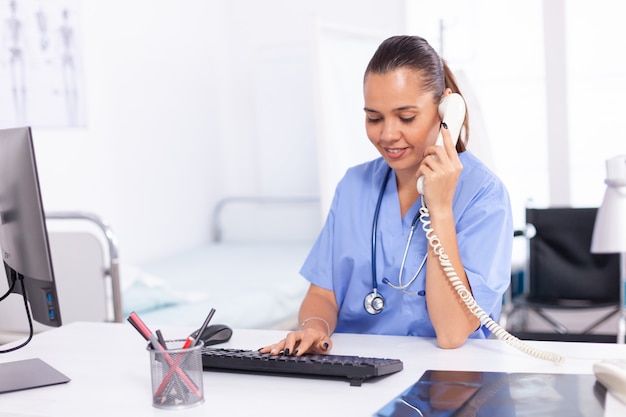 Enfermeira ligando para o paciente da recepção do hospital para falar sobre o diagnóstico. médico de saúde sentado na mesa usando o computador na clínica moderna, olhando para o monitor.