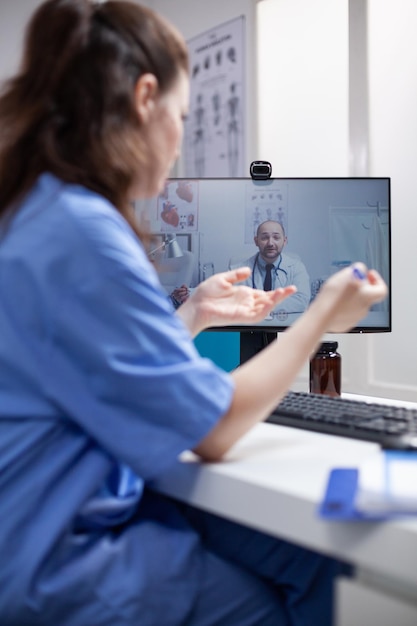 Enfermeira na mesa em videochamada de telemedicina comunicando-se com médico especialista em consultório médico profissional. Médico tendo videoconferência on-line na Internet com um jovem colega