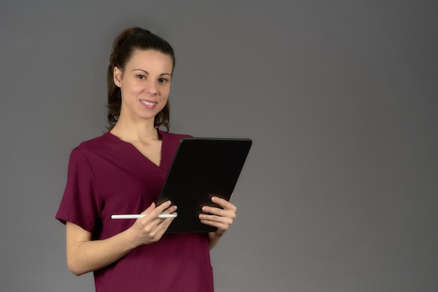 Enfermeira sorridente de uniforme roxo olha para a câmera com relatório de tablet digital em fundo cinza