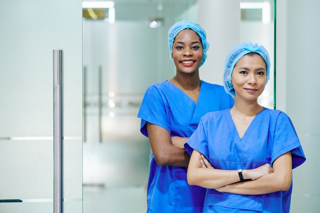 Enfermeiras médicas multiétnicas e sorridentes, de uniforme e bonés descartáveis, paradas no corredor do hospital