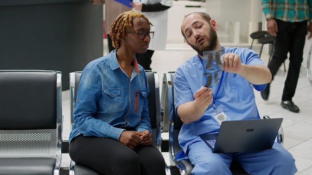 Enfermeiro masculino mostrando resultados de radiografia de ossos para paciente sentado no saguão de recepção do hospital. especialista e jovem olhando para a varredura de raio-x para ver o diagnóstico de saúde na área de espera.