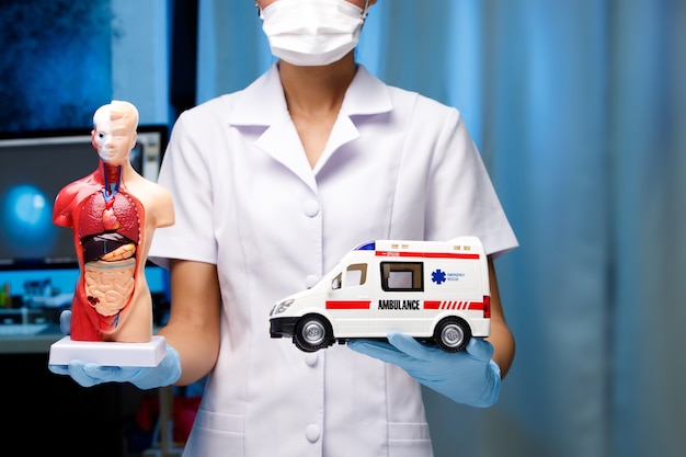 La enfermera abre con dos manos y muestra la figura de la anatomía humana y el vehículo de ambulancia en el cartel de la palma con la sala de fondo de tono azul del hospital