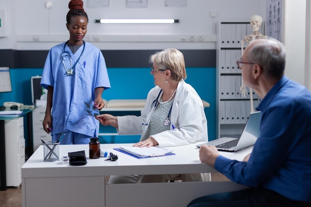 Enfermera afroamericana dando radiografías al médico para consultar con pacientes enfermos en el gabinete. Médico que recibe los resultados del escaneo de rayos X del asistente para explicar el diagnóstico al anciano.