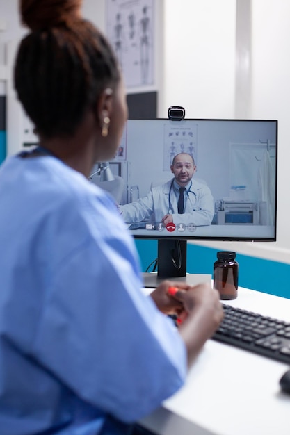 Enfermera afroamericana en el escritorio en una videollamada comunicándose con un colega médico en un consultorio médico profesional. Médico especialista en atención médica que tiene una reunión de equipo en línea con un compañero de trabajo