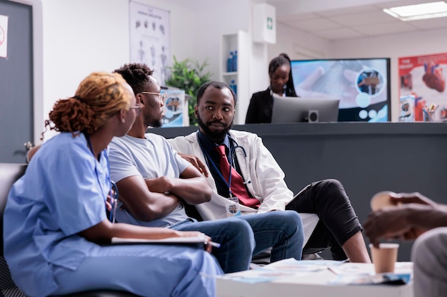 Enfermera afroamericana y médico hablando con el paciente, consultando con un adulto joven en la sala de espera del vestíbulo del hospital. Equipo médico ayudando al hombre con tratamiento y medicamentos en la sala de espera.