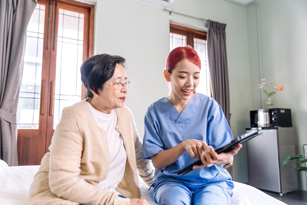 Enfermera asiática que usa batas informa el estado de salud de la mujer asiática mayor con tableta digital en el dormitorio Visita del cuidador en el hogar Concepto de cuidado de la salud en el hogar y hogar de ancianos