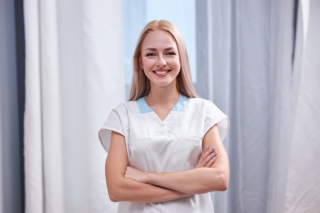Enfermera en bata blanca de pie con los brazos cruzados, mujer médico practicante posando y sonriendo, concepto de medicina y atención médica