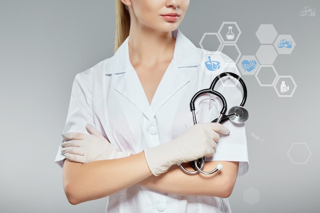 Enfermera con bata médica blanca y sosteniendo estetoscopio en fondo gris, concepto de farmacología y atención médica, chica en uniforme blanco, de cerca, media cara