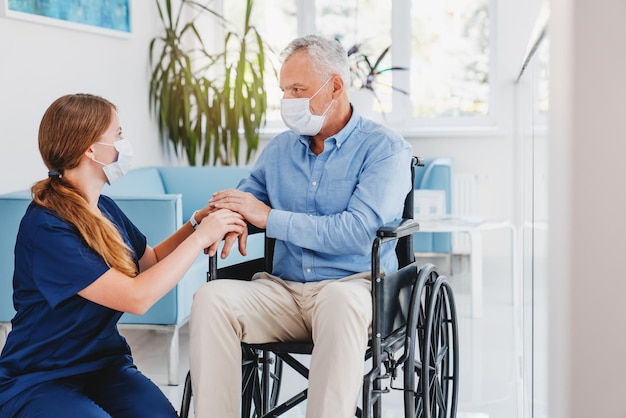 Enfermera caucásica apoyando a un anciano en silla de ruedas que lleva máscaras médicas de protección quirúrgica en el vestíbulo del hospital Concepto coronavirus covid 19