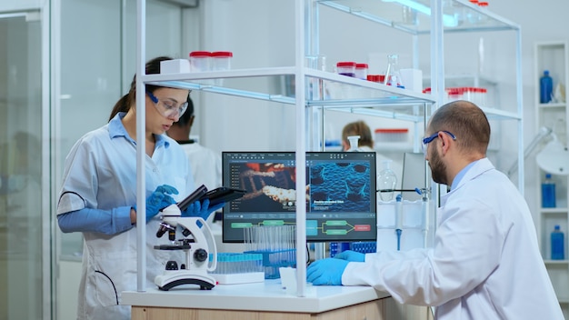 Enfermera científica tomando notas sobre la tableta en el moderno laboratorio equipado. Equipo multiétnico que examina la evolución de la vacuna utilizando alta tecnología para la investigación en el desarrollo de tratamientos contra el virus covid19