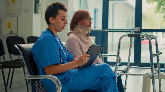 Enfermera consultando al paciente con collarín de cuello y andador, tomando notas sobre lesiones por accidente en la tableta. Haciendo una visita de control con una mujer que usa espuma cervical y deterioro.