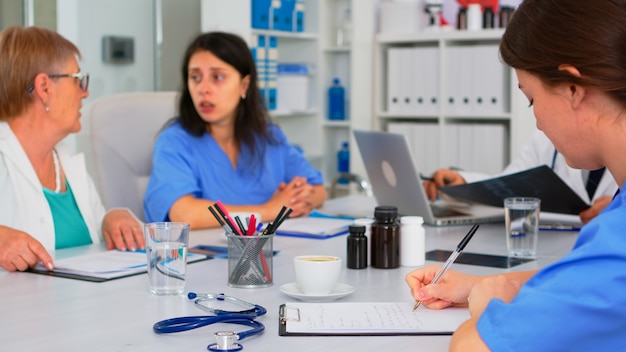 Enfermera escribiendo en el portapapeles mientras los trabajadores en equipo profesionales que tienen una reunión médica discutiendo en segundo plano en la oficina de intercambio de ideas. Médicos profesionales que examinan los síntomas del paciente en la sala de reuniones.