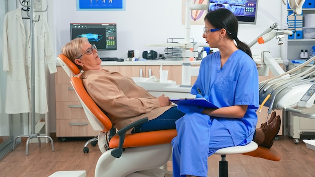 Enfermera explicando el procedimiento médico al médico dentista de espera del paciente senior en la sala de estomatología. Asistente tomando notas en el portapapeles hablando con anciana preparándose para examen estomatológico