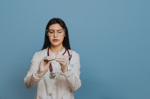 Enfermera con gafas, vestida con una bata médica, revisando el tubo de ensayo, concentrándose en estudiar virus, descubriendo la vacuna. Concepto de virus Covid-19. Sobre fondo azul con espacio de copia