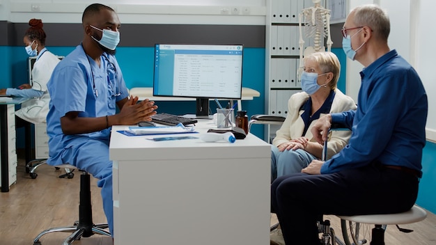 Enfermera hablando con una mujer con discapacidad y su marido en una cita de consulta durante la pandemia del coronavirus. Especialista que examina al paciente con discapacidad en silla de ruedas en la visita de control.