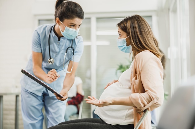 Enfermera hablando con una mujer embarazada preocupada y analizando el informe médico antes de la vacuna covid-19 en la sala de espera del hospital.