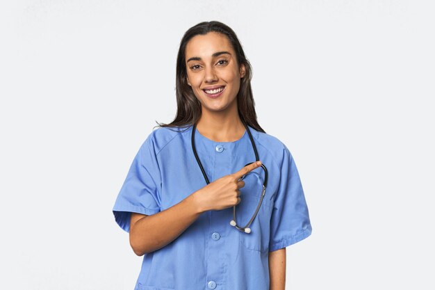 Foto enfermera hispana en uniforme con estetoscopio sonriendo y señalando a un lado mostrando algo