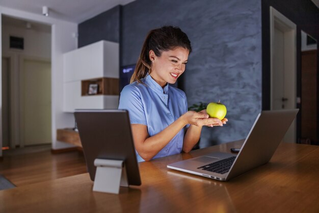 Enfermera joven amistosa sonriente que se sienta en casa y que sostiene la manzana. Ella está dando consejos para una vida sana a través de Internet.