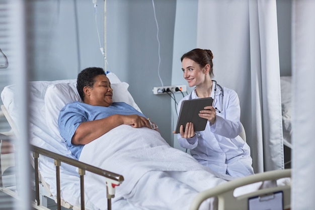 Enfermera joven sonriente que muestra la tableta digital a la mujer mayor en el cuarto de hospital