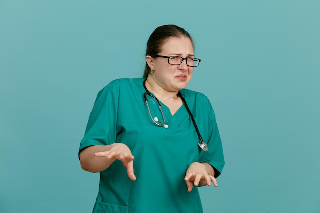 Enfermera joven en uniforme médico con estetoscopio alrededor del cuello mirando a la cámara con expresión disgustada haciendo gesto de defensa con las manos sobre fondo azul.