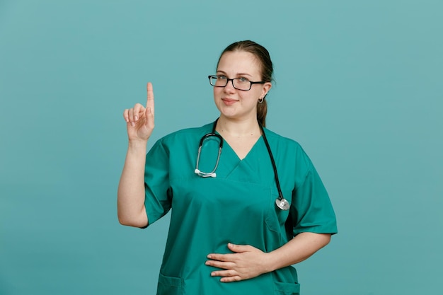 Enfermera joven en uniforme médico con estetoscopio alrededor del cuello mirando a la cámara sonriendo confiada mostrando el dedo índice teniendo una buena idea de pie sobre fondo azul