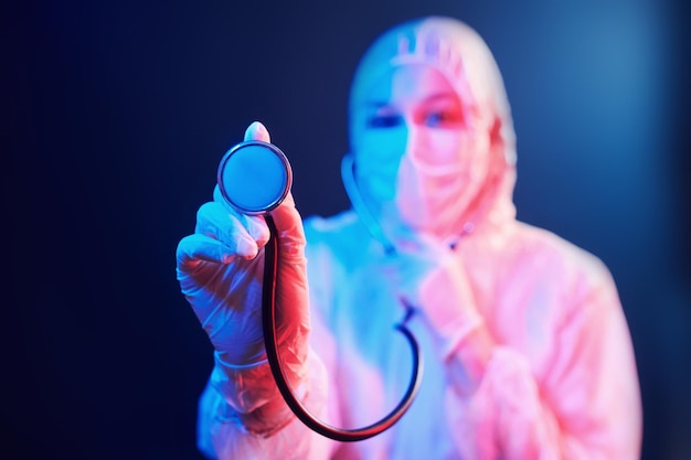 Enfermera con máscara y uniforme blanco y con estetoscopio de pie en una habitación iluminada con neón