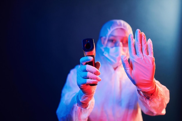Enfermera con máscara y uniforme blanco de pie en una habitación iluminada con neón y sosteniendo un termómetro infrarrojo Detener la propagación del coronavirus