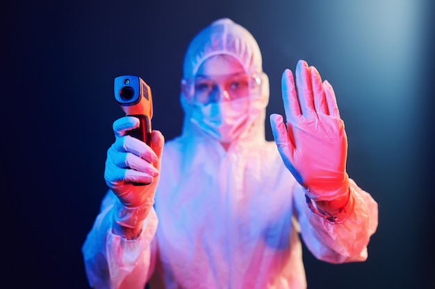 Enfermera en máscara y uniforme blanco de pie en la habitación iluminada de neón y sosteniendo el termómetro infrarrojo. Detener la propagación del coronavirus