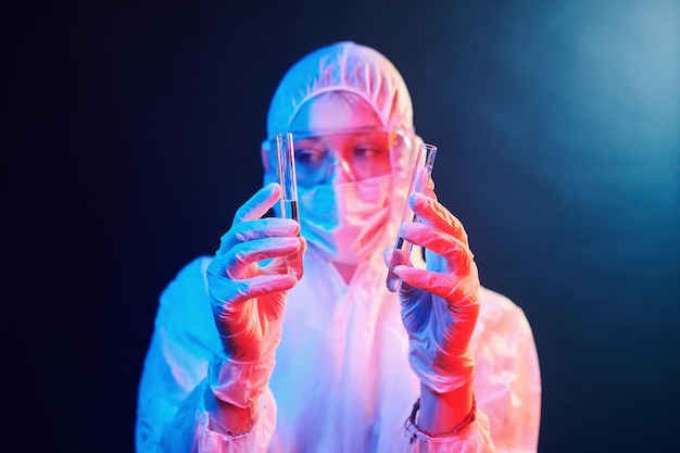 Enfermera con máscara y uniforme blanco de pie en una habitación iluminada con neón y sosteniendo tubos con muestras