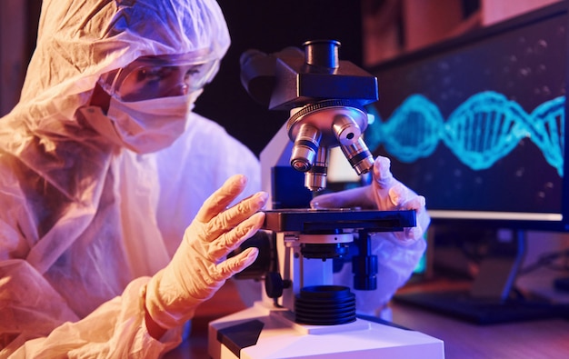 Enfermera con máscara y uniforme blanco sentada en un laboratorio iluminado de neón con computadora, microscopio y equipo médico en busca de la vacuna contra el coronavirus