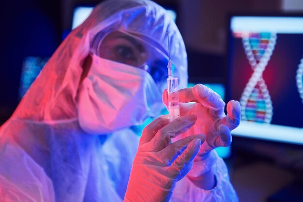 Enfermera con máscara y uniforme blanco sosteniendo una jeringa y sentada en un laboratorio iluminado con neón con computadora y equipo médico en busca de la vacuna contra el coronavirus