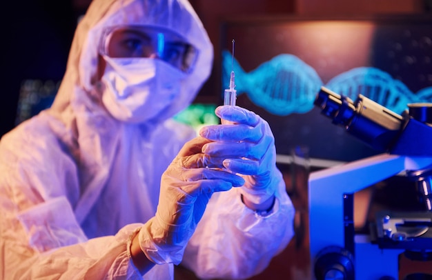 Enfermera con máscara y uniforme blanco, sosteniendo una jeringa y sentada en un laboratorio iluminado de neón con computadora y equipo médico en busca de la vacuna contra el coronavirus