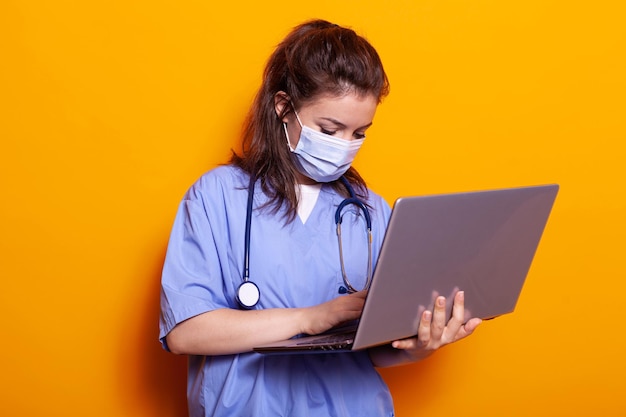 Enfermera con mascarilla mirando la laptop y usando uniforme y estetoscopio. Asistente médico que usa computadora moderna durante una pandemia, persona de pie sobre un fondo aislado.