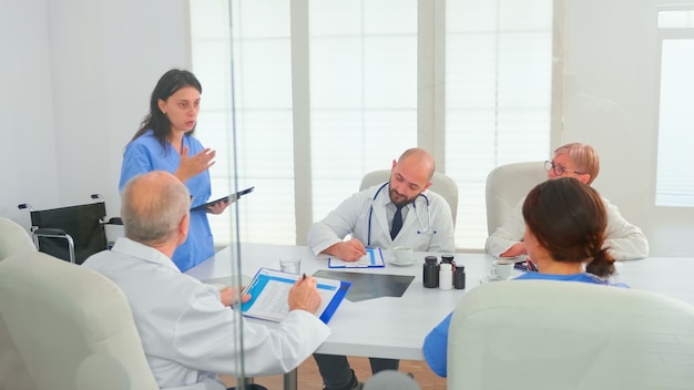 Enfermera médica joven que usa la tableta en la sala de conferencias del hospital con compañeros de trabajo que explican el desarrollo de la enfermedad. Terapeuta experto de la clínica hablando con colegas sobre la enfermedad, profesional de la medicina