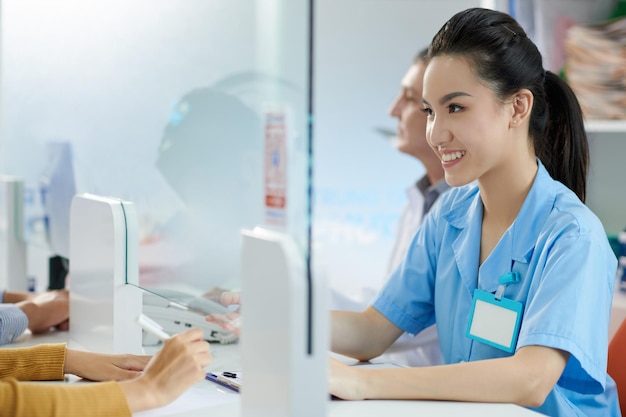 Enfermera médica joven sonriente que trabaja en la recepción del hospital, hablando con pacientes y llenando documentos