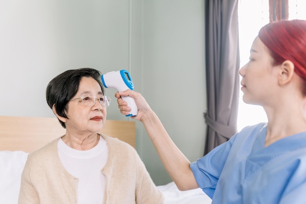 Una enfermera mide la temperatura de una anciana asiática usando una pistola de termómetro de frente infrarroja en su hogar. Visita del cuidador en el hogar. Concepto de cuidado de la salud y hogar de ancianos.