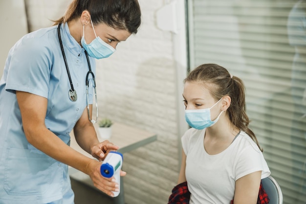 Enfermera midiendo la temperatura de una linda adolescente en la sala de espera durante la pandemia del coronavirus.