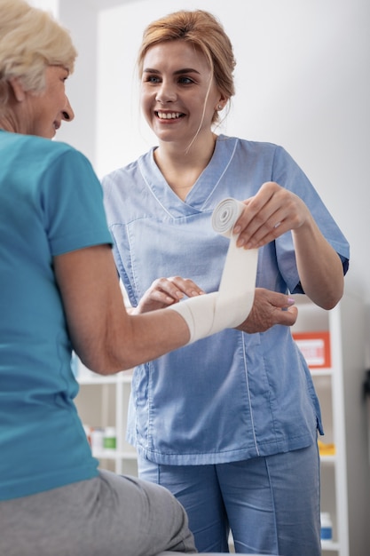 Enfermera profesional especializada. Bonita mujer guapa sonriendo mientras habla con su paciente durante el vendaje