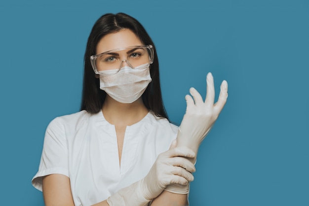 Una enfermera profesional con gafas protectoras, una máscara quirúrgica y una bata médica usa sus guantes.