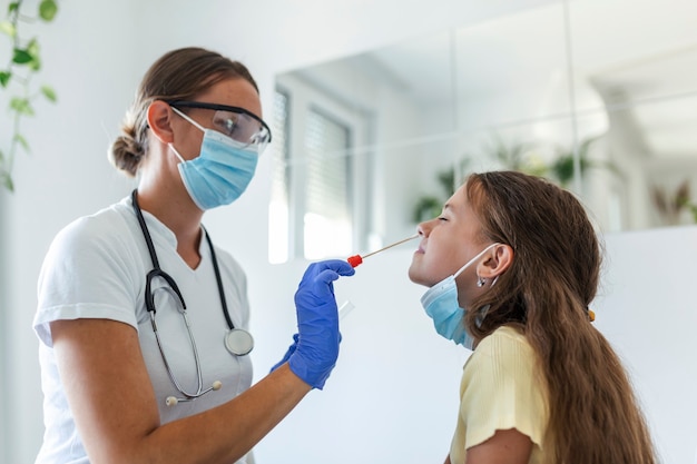 Enfermera realizando una prueba de frotis nasal en un niño pequeño. Niña pasando por una prueba de PCR debido a la pandemia de COVID-19. Doctora con hisopo de algodón mientras prueba de PCR niña pequeña