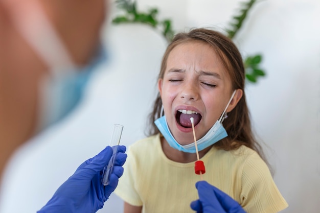 Enfermera realizando una prueba de hisopado bucal en un niño pequeño. Niña pasando por una prueba de PCR debido a la pandemia de COVID-19. Doctora con hisopo de algodón mientras prueba de PCR niña pequeña