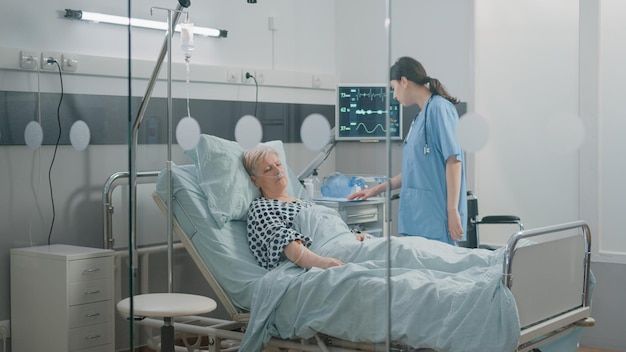 Enfermera revisando a un anciano dormido en la cama para recibir tratamiento médico. Asistente médico mirando el monitor de frecuencia cardíaca para medir el pulso. Especialista y pensionista en sala de hospital