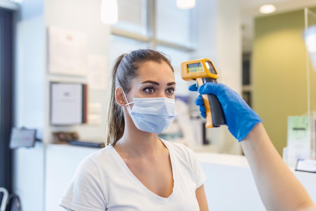 Enfermera revisando la temperatura de la mujer con un termómetro infrarrojo en el mostrador La mujer lleva una mascarilla protectora Están en el hospital durante la pandemia