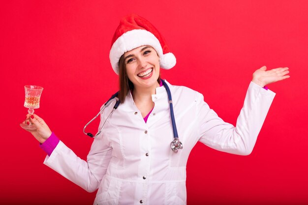 Una enfermera con un sombrero de Papá Noel sostiene una copa de champán feliz año nuevo con un fondo rojo