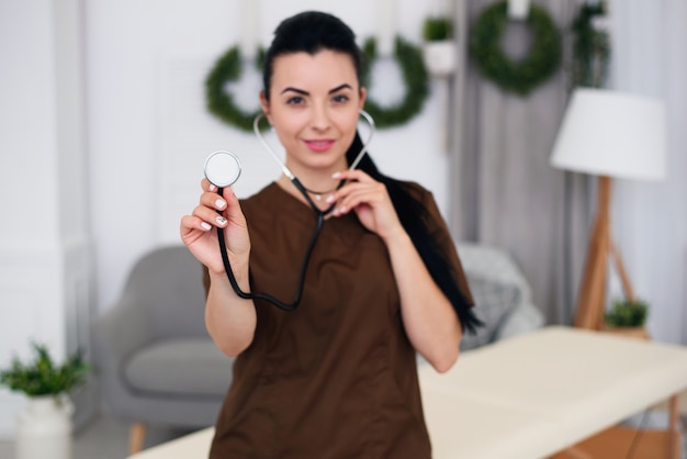 Enfermera sonriente hermosa que lleva el traje médico marrón con el estetoscopio a disposición en oficina médica moderna. Concepto de salud y farmacología, chica en uniforme.