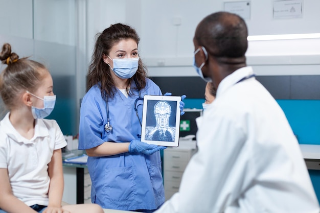 Enfermera terapeuta con mascarilla de protección contra el coronavirus sosteniendo una tableta con radiografía del paciente en la pantalla. Médico pediatra afroamericano que trabaja en la oficina del hospital