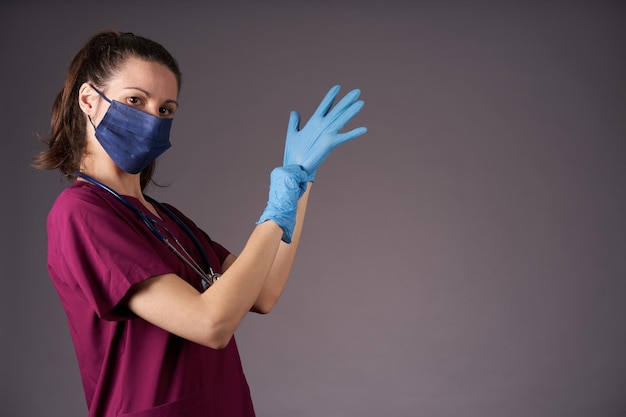 Enfermera con uniforme morado poniéndose guantes médicos de látex mirando a la cámara con fondo gris