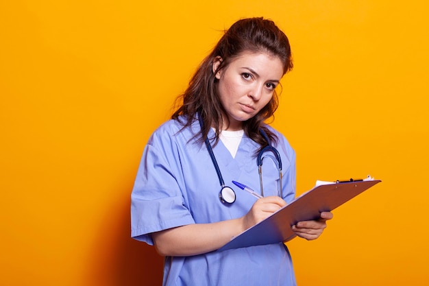 Enfermera con uniforme tomando notas en los archivos del portapapeles, de pie sobre un fondo aislado. Asistente médico escribiendo en documentos para tener información de atención médica, mirando la cámara