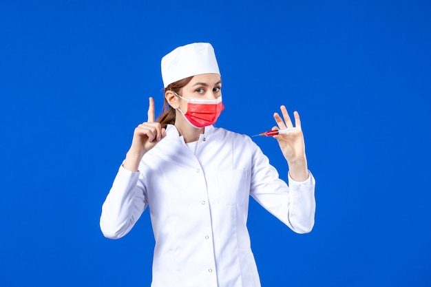Enfermera de vista frontal en traje médico blanco con máscara roja e inyección en sus manos en azul
